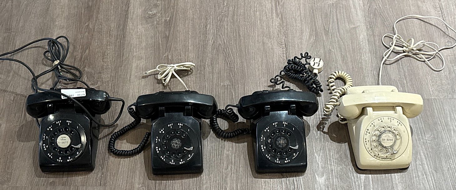 Four rotary desk telephones in a row, three black and one ivory, seen from above with the handsets on top